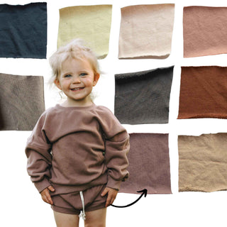 Assortment of different swatches for our shorties. They are made by My Mini Explorer in their family owned and operated workshop. Colors include atlantic, mellow, parchment, rose clay, mushroom, graphite, tawny, lilac, and barley. The little girl in the picture is wearing a lilac crewneck with lilac shorites.